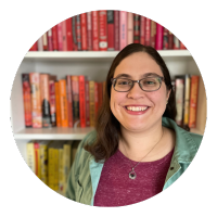 Erin Madison, a light-skinned woman with brown glasses, brown, wavy shoulder-length hair, is in front of a colorful bookshelf that is full of books. Erin is smiling widely and has dimples. She is wearing a dark rose-colored sweater and a teal jacket and has a silver necklace that has a pendant with a purple gem.